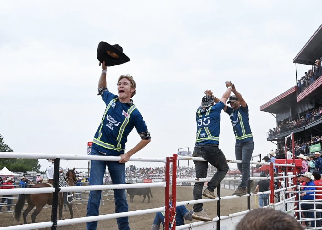 Scene at a rodeo taken from the crowd through the railings to the arena. A tall red stadium is on the right hand side, and three cowboys are hanging onto the gates. The front cowboy has his hat in the air and the back two cowboys are giving each other a high-five