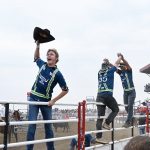 Scene at a rodeo taken from the crowd through the railings to the arena. A tall red stadium is on the right hand side, and three cowboys are hanging onto the gates. The front cowboy has his hat in the air and the back two cowboys are giving each other a high-five