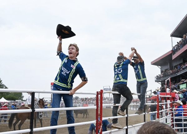 Scene at a rodeo taken from the crowd through the railings to the arena. A tall red stadium is on the right hand side, and three cowboys are hanging onto the gates. The front cowboy has his hat in the air and the back two cowboys are giving each other a high-five