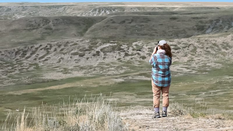 A large valley covered in grasslands stretches across the frame. Small strip of pale blue sky above. A lade in baseball hat, plaid shirt and brown trousers stands on a cliff in foreground taking a photo of the valley