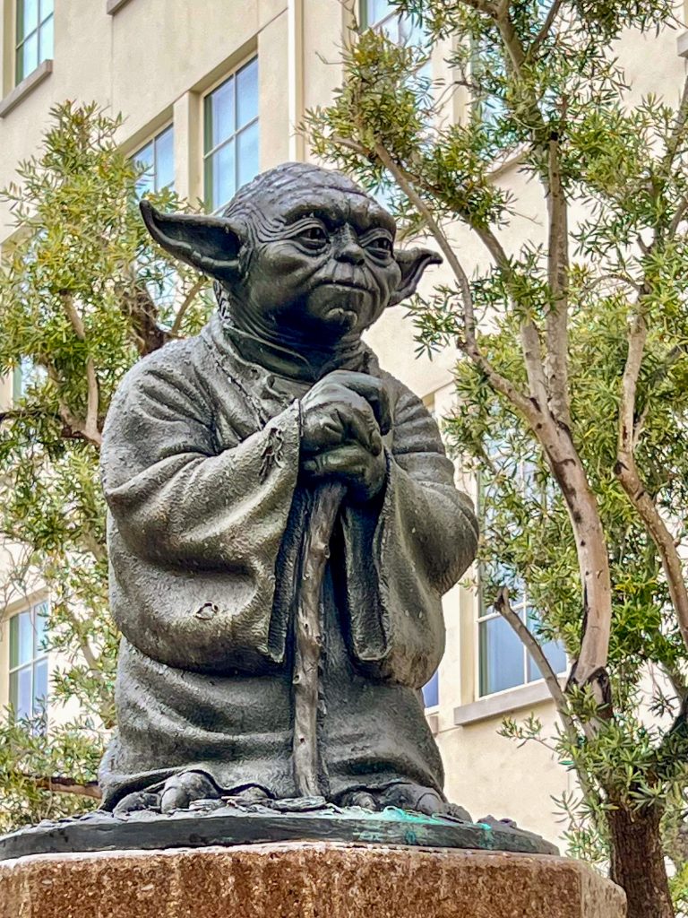Statue of Star Wars character, Yoda, in bronze sitting in front of office building behind with trees.