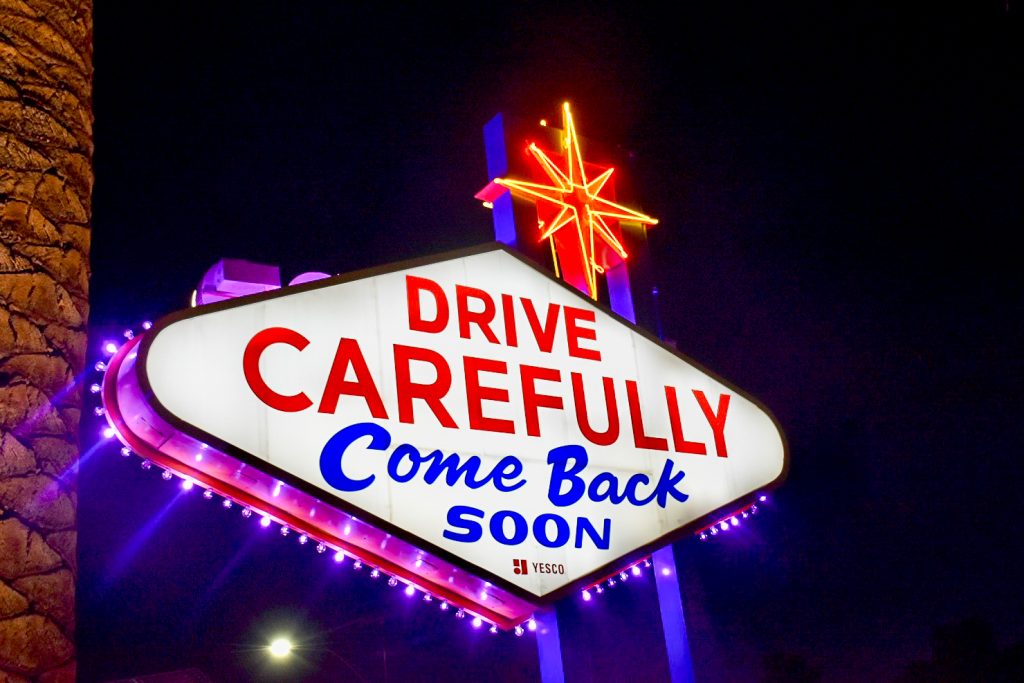 Sky at night, edge of palm tree to the left hand side. Illuminated sign in the bottom left of photo saying Drive Carefully, Come Back Soon. Red and blue lettering, pink and blue neon lights. Orange neon star to the top right
