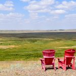 Two red Adirondack chairs facing away from the camera (bottom right hand side) and over a valley expanse of grasslands. Pale blue sky above with white fluffy clouds
