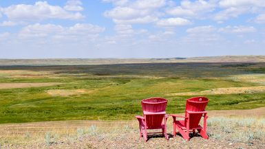 Two red Adirondack chairs facing away from the camera (bottom right hand side) and over a valley expanse of grasslands. Pale blue sky above with white fluffy clouds