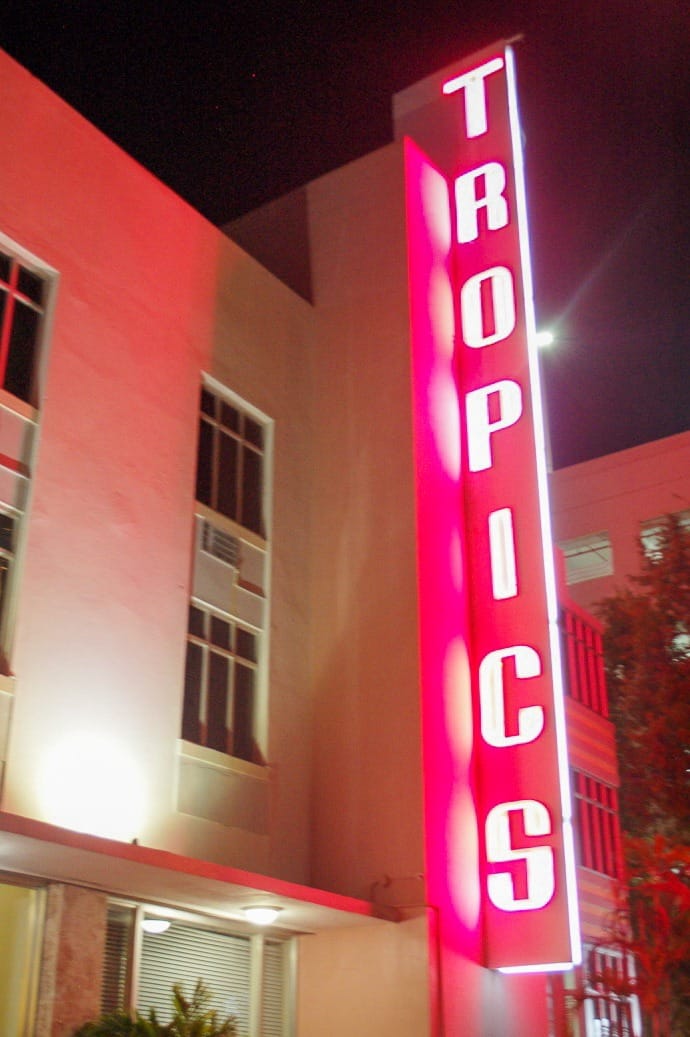 Portrait shape. Photo taken at night of a white art deco building with illuminated vertical pink neon sign saying Tropics.