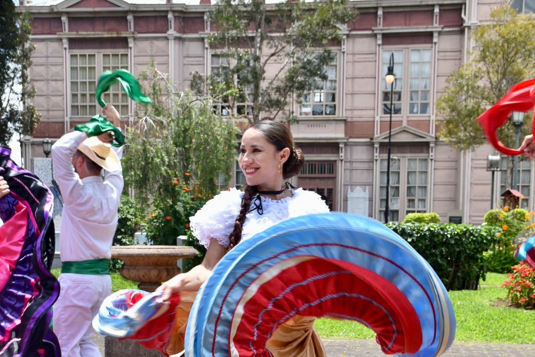 Smiling lady with brown hair scraped back in a white blouse is smiling to the left and twirling a blue, red and gold skirt in a circle towards the camera. Other dancers behind her and a pale pink building the windows in the background