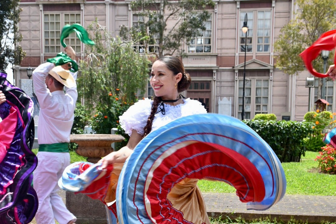 Smiling lady with brown hair scraped back in a white blouse is smiling to the left and twirling a blue, red and gold skirt in a circle towards the camera. Other dancers behind her and a pale pink building the windows in the background