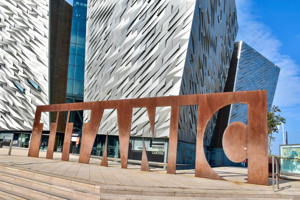 A bronze metal statue spelling the word TITANIC sitting outside a modern geometric building with silver walls.