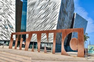 A bronze metal statue spelling the word TITANIC sitting outside a modern geometric building with silver walls.