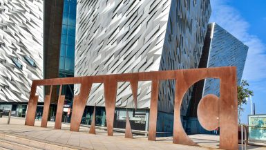 A bronze metal statue spelling the word TITANIC sitting outside a modern geometric building with silver walls.