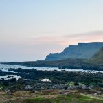 A landscape scene of dark rocks leading into the sea in the foreground, being a small strip of grass. A headland cliff can be seen behind with the pink of a sun rise against an otherwise grey sky