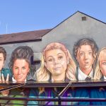 Mural on the side of a grey building with blue sky behind. The Mural features 5 faces - a boy on the left, then girl with brown hair, girl with blonde hair, girls with brown hair and girl with blond hair. They are wearing school uniform. There is sun shade attached to the wall of the building which cuts across the bottom of the mural