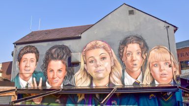 Mural on the side of a grey building with blue sky behind. The Mural features 5 faces - a boy on the left, then girl with brown hair, girl with blonde hair, girls with brown hair and girl with blond hair. They are wearing school uniform. There is sun shade attached to the wall of the building which cuts across the bottom of the mural