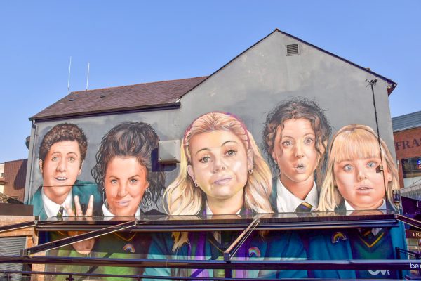 Mural on the side of a grey building with blue sky behind. The Mural features 5 faces - a boy on the left, then girl with brown hair, girl with blonde hair, girls with brown hair and girl with blond hair. They are wearing school uniform. There is sun shade attached to the wall of the building which cuts across the bottom of the mural