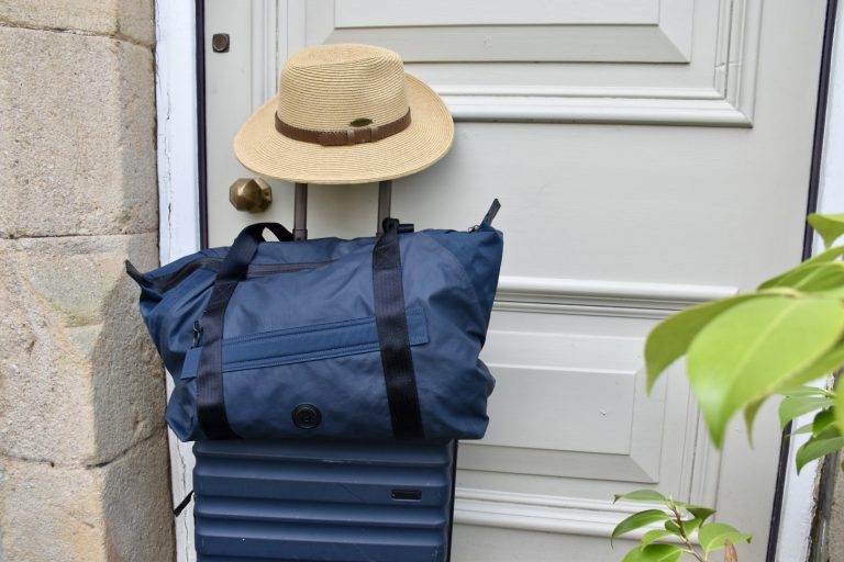 Navy blue roller suitcase, with a navy blue soft bag sitting on top of it parked outside a wooden front door painted pale green. A Panama straw hat sits on the handles of the suitcase. Leaves of plant visible on right hand side.