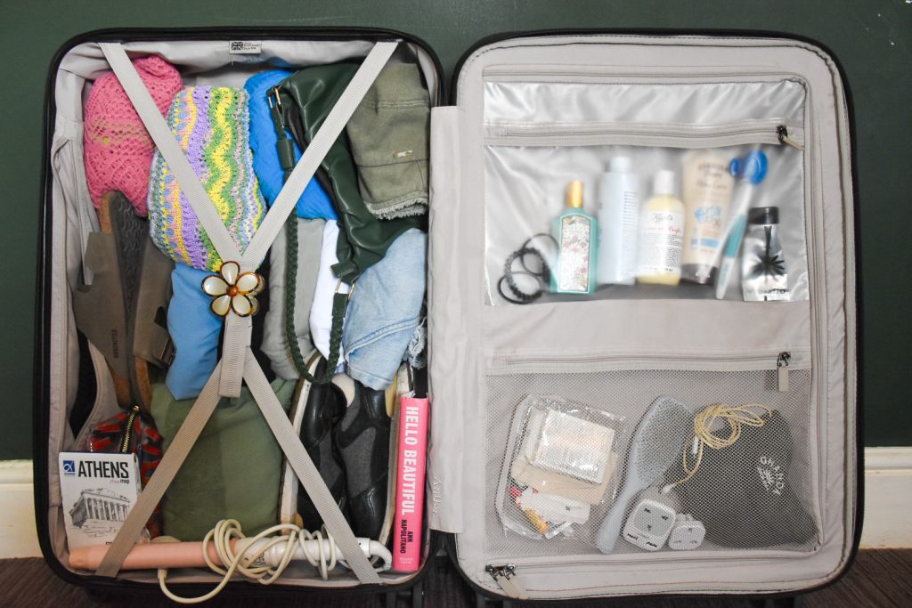 Frame is filled with the inside contents of an open suitcase. On the left hand side you can see folded clothes, trainers and other packed items held in place by crossed elastic straps. On the right hand side there is a zipped cover across the contents with pockets filled with toiletries