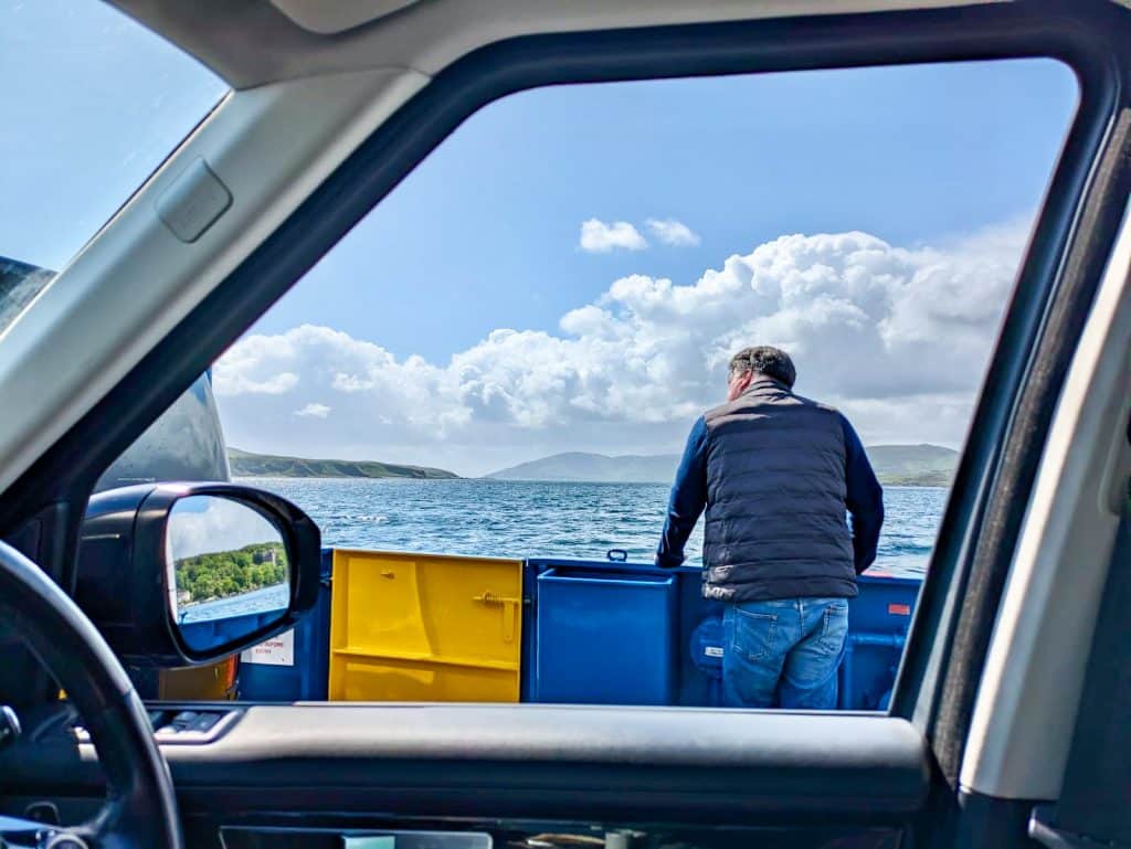 Photo taken from the inside of a car, fat bloke looking out. Through the windows its a man in jeans and dark jacket looking out over a body of water with hills, sky and clouds in the background