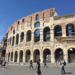 The outside curve of a tiered amphitheatre, three stories high with arches on each layer. There are people walking around the outside of the structure