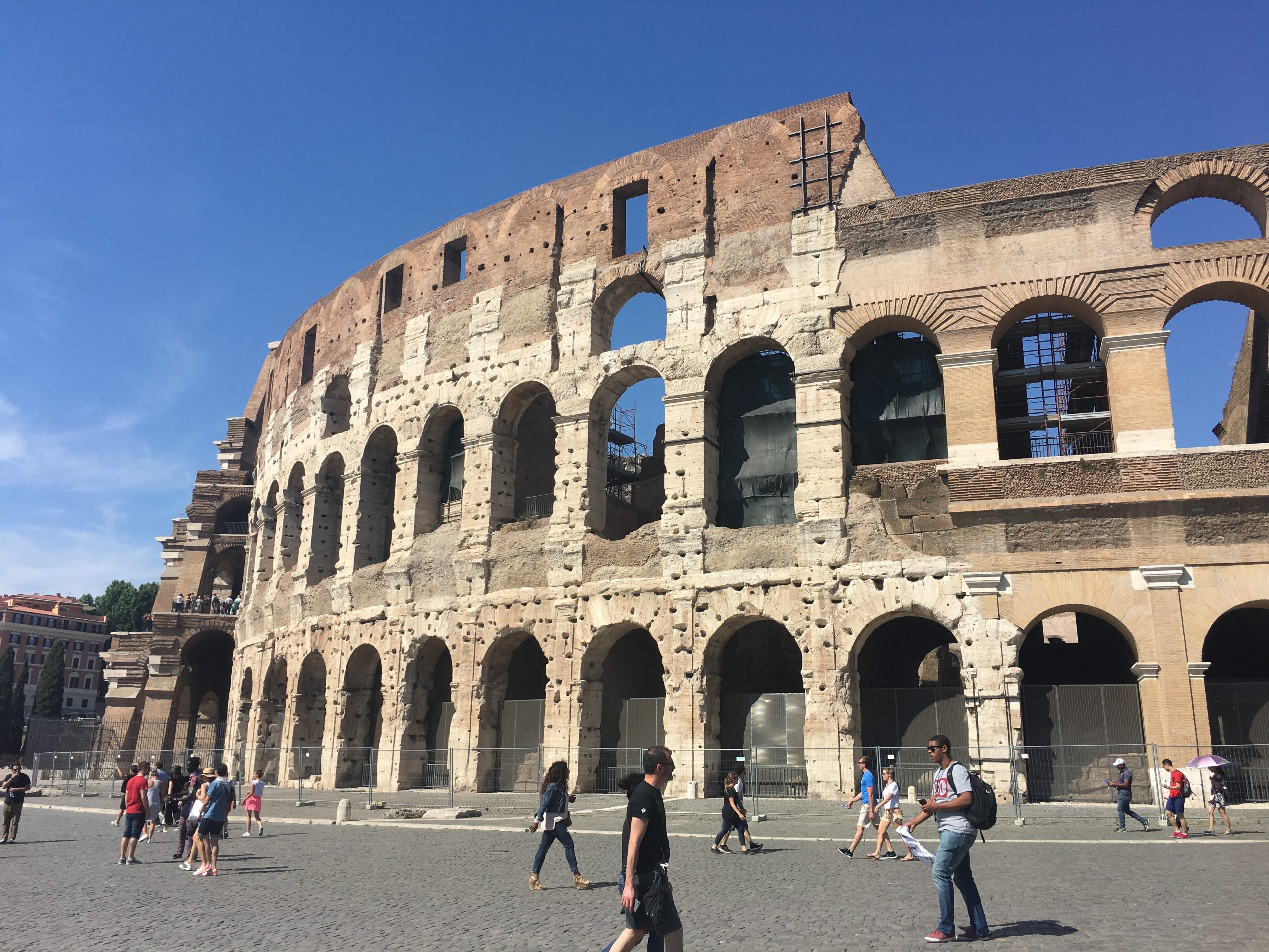 The outside curve of a tiered amphitheatre, three stories high with arches on each layer. There are people walking around the outside of the structure