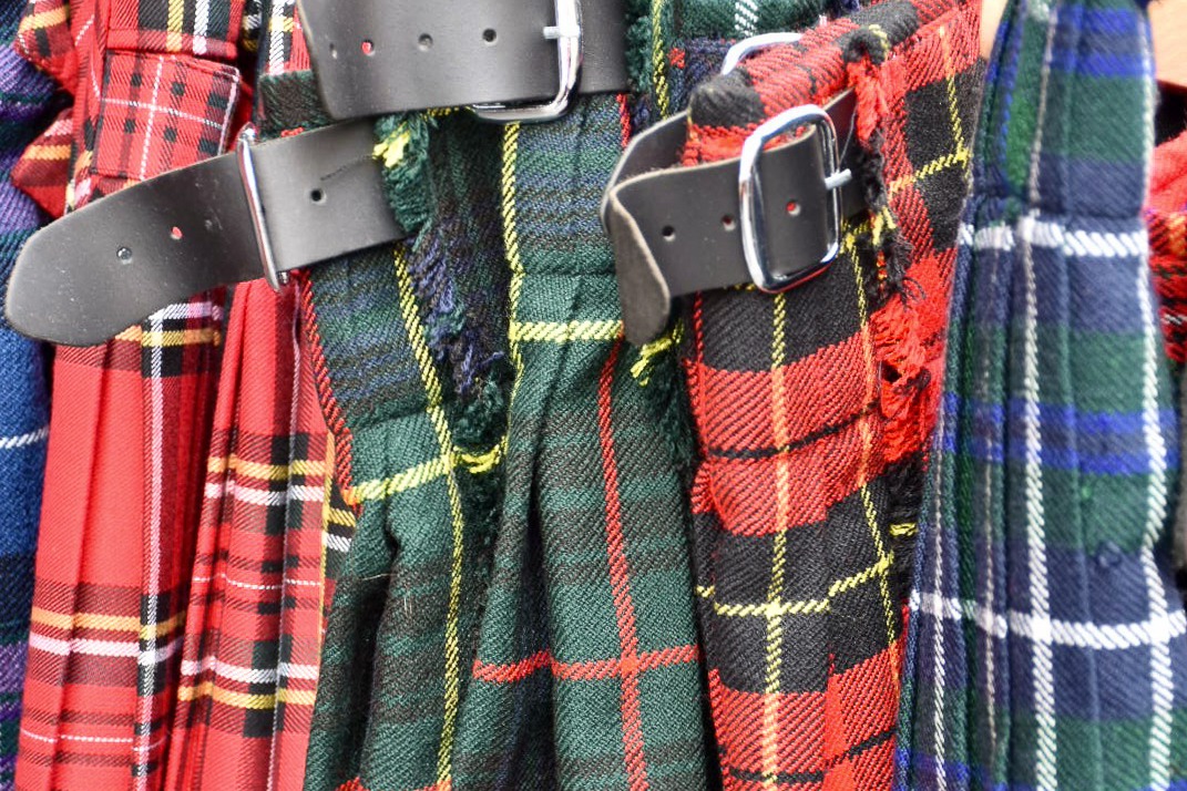 Close up of a row of tartan kilts, mixed colours and plaid. Leather buckles visible at the top