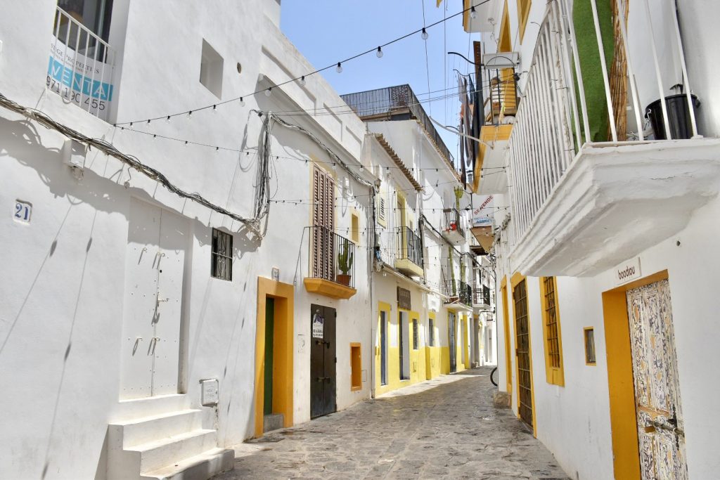 Street of white buildings on either side of a narrow paved street. Strings of lightbulbs across the street. Houses have doorways trimmed with mustard yellow paint, and upstairs iron balconies