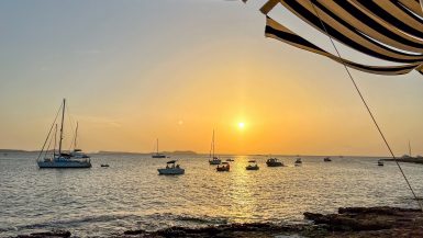 A sunset landscape taken from the shoreline over the water. There are sailboats on the water. You can see a stripe awning at the top of the frame