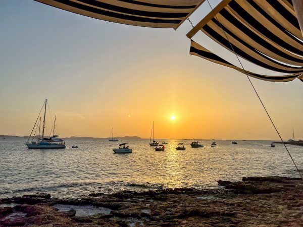 A sunset landscape taken from the shoreline over the water. There are sailboats on the water. You can see a stripe awning at the top of the frame