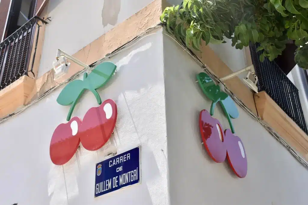 View of a street corner - white walls. Blue sign on the left saying 'Career de Guillem de Montgri'. It has two cherries with green leaves as a motif on both the left and right sides of the corner