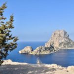Photo taken from a pale rocky cliff out to sea which is flat a blue. A white rocky outcrop stands just off the shore. Blue sky behind. Pine tree to the left.
