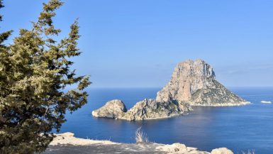 Photo taken from a pale rocky cliff out to sea which is flat a blue. A white rocky outcrop stands just off the shore. Blue sky behind. Pine tree to the left.