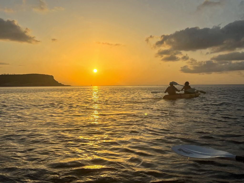 Sunset shot taken from a kayak on the water. You can see an oar in the bottom left. A double kayak is making its way to the middle right of the frame. Sun is setting left of centre. a couple of grey clouds