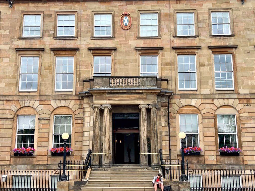 A large stone building fills the frame with three stories of rectangular windows. There are window boxes on the bottom windows with pink flowers. A set of steps leads up to an impressive doorway with columns on either side. A figure sits on the bottom step