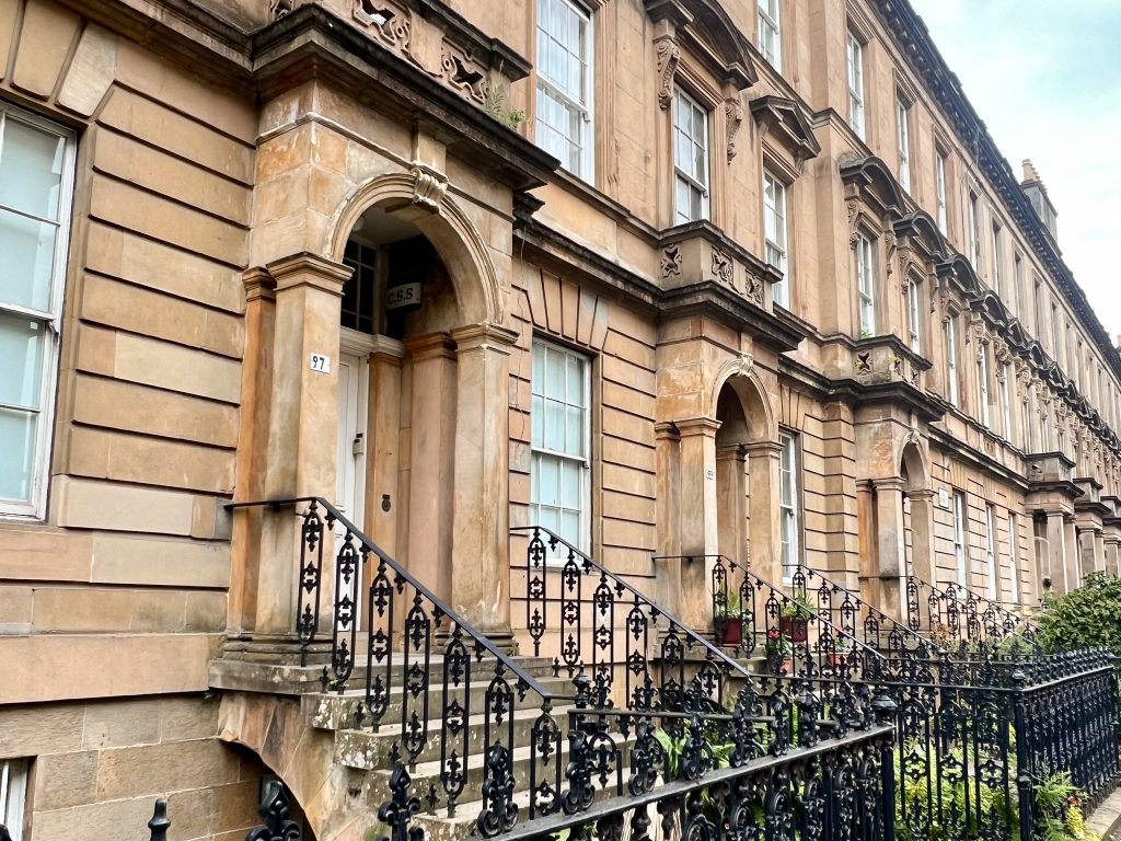 Row of brown sandstone houses reaching from left hand side to the middle right. The houses have stairs up to the front doors with black iron railings.