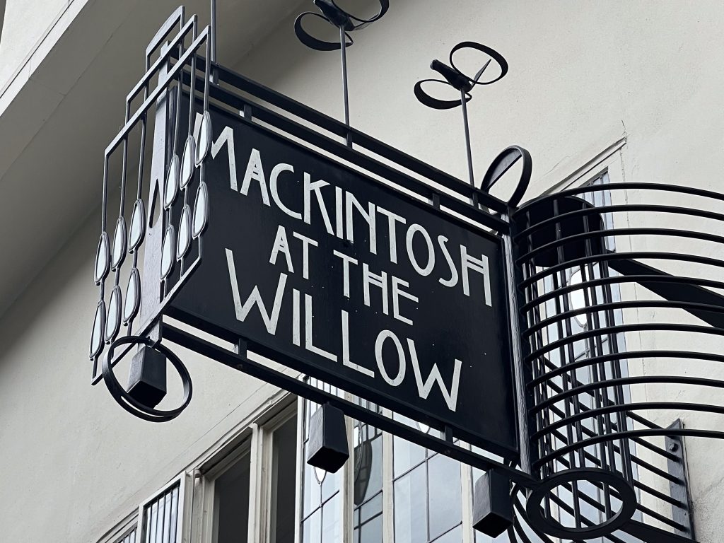 Black iron sign attached to a white building. The sign is Art Nouveau style and stays Mackintosh at the Willow on it in white