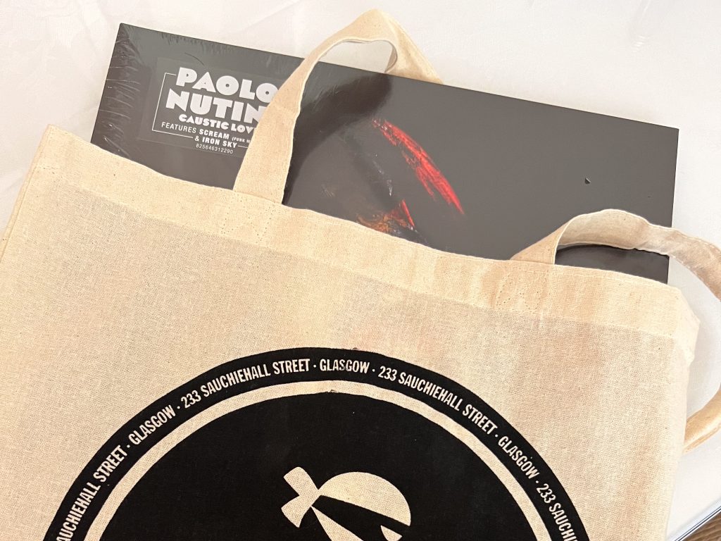 A cream cotton bag sits on a white surface. The bag features a black design advertising a shop on 233 Sauchiehall Street. A vinyl record can be seen coming out of the top of the bag, by Paolo Nutini (Caustic Love)