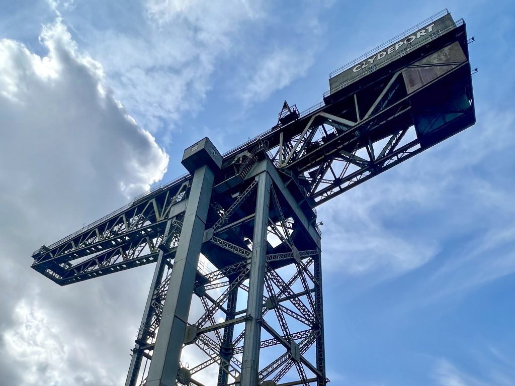 Blue sky with whispy white cloud to the left. From the bottom centre of the frame rises a large metal crane with two arms. The one on the right has the word Clydeport printed in white
