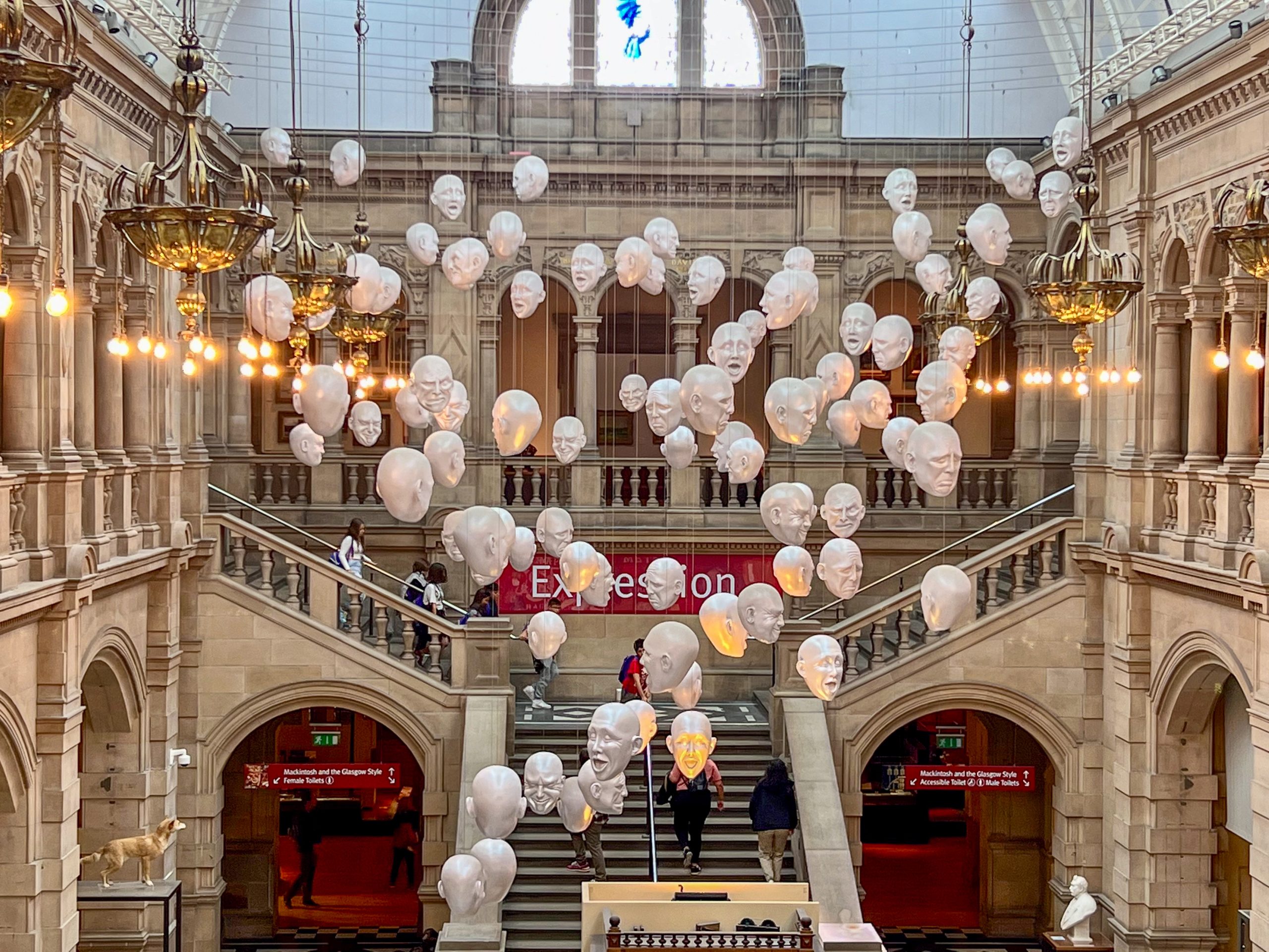 Inside an old stone building, looks like a museum with grand stairway leaving to the lower floor. There are around 50 white heads hanging from the ceiling in an art display