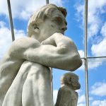 White marble statue of a man in a thinking pose, with a monkey sitting next to him but looking out of the window behind. The shot is taken upwards through panes of glass to a blue sky with white clouds