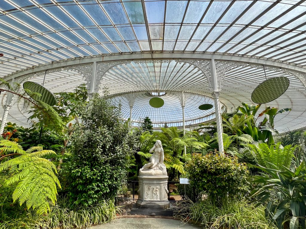 Inside the curved dome of a glasshouse with tropical plants at the bottom and a white statue of a lady sits in the middle of the frame