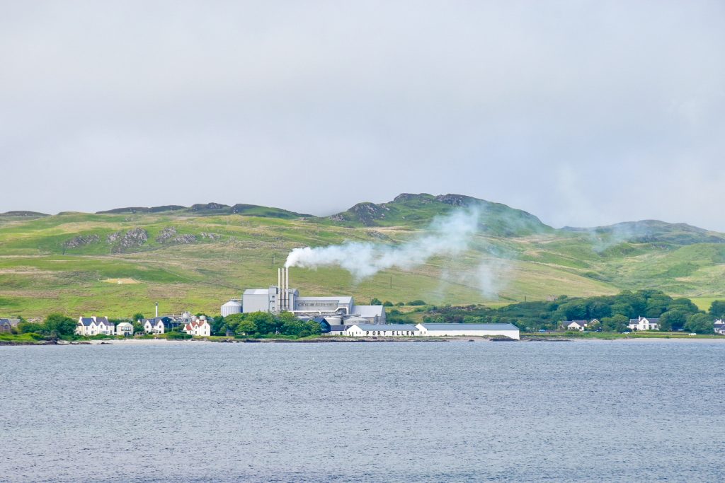 The bottom third of the frame shows a flat blue-grey sea, the top third a pale grey sky. The middle third shows a row of green hills with some white buildings and a factory in front. There is smoke coming from 3 silver factory chimneys. The long thin white building at the front of the factory says Port Ellen on the front in large black letters