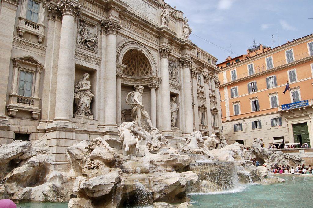 Large white marble carvings of a water fountain above turquoise water. Orange building in the background