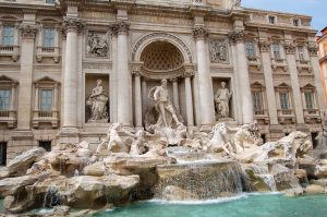 Large baroque marble fountain facade featuring a central figure above a number of horses. Turquoise water in the bottom right of frame around fountain