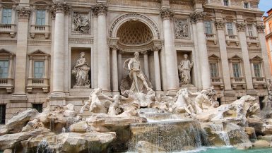 Large baroque marble fountain facade featuring a central figure above a number of horses. Turquoise water in the bottom right of frame around fountain
