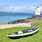 Landscape of two square lighthouse towers standing on rocks overlooking a body of water with strip of green land beyond. In the foreground is a sandy beach, and grassy bank. There is an inflatable kayak (black and green) in the foreground