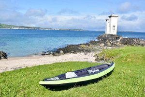 Landscape of two square lighthouse towers standing on rocks overlooking a body of water with strip of green land beyond. In the foreground is a sandy beach, and grassy bank. There is an inflatable kayak (black and green) in the foreground