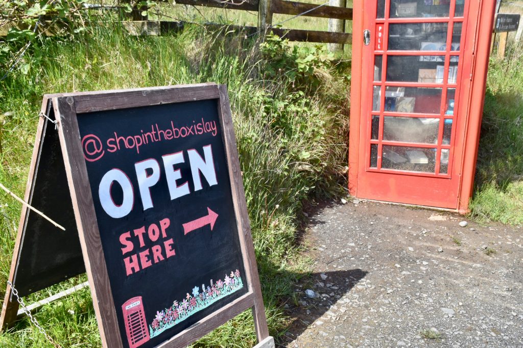 A red telephone box stands at the top right of the frame. A wooden sign stands in the bottom left of the frame. It is advertising the Shop in the Box, Islay. Open. Stop here with an arrow towards the phone box. Grass behind.