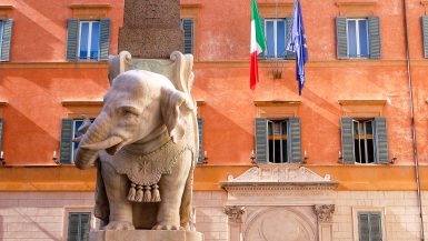 Beautiful carving of an elephant looking to the left in white marble. Obelisk behind. Ocre coloured building behind. Features two flags (Italy and Europe) hand outside the building.