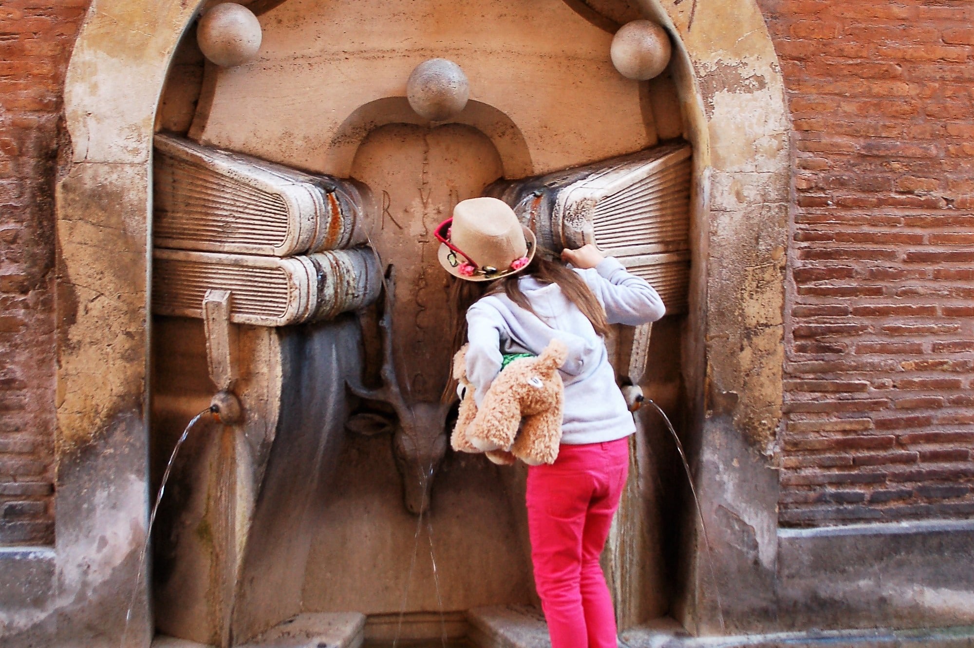 Young girl (pink trousers, grey hoody, straw hat and teddy under arm) drinks from a stone fountain with a book design