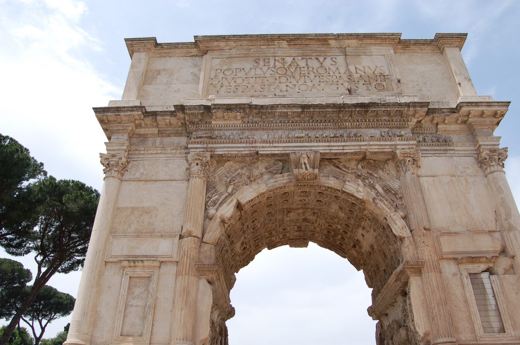 Upwards view of a large marble column with Latin words at the top. Pale grey sky behind. Cypress tree to the left
