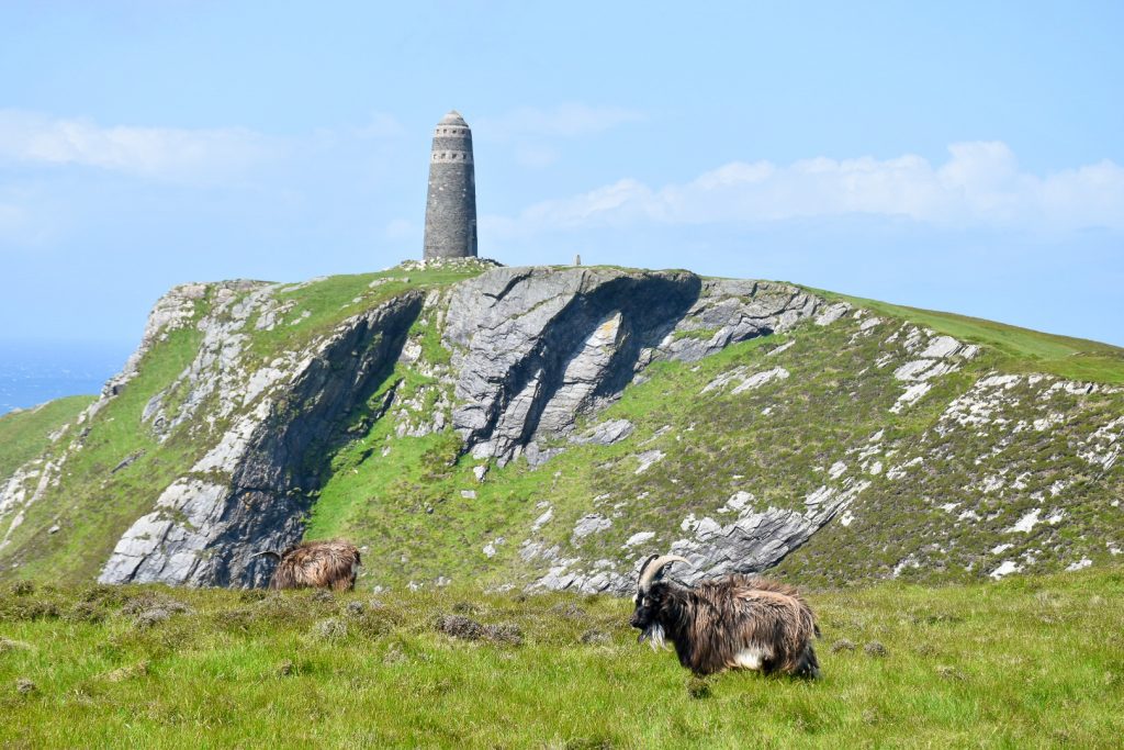 A stone monument stands on top of a green rocky cliff. In the foreground a horned brown goat is making its way from the bottom right to left of the frame. Pale blue sky above.